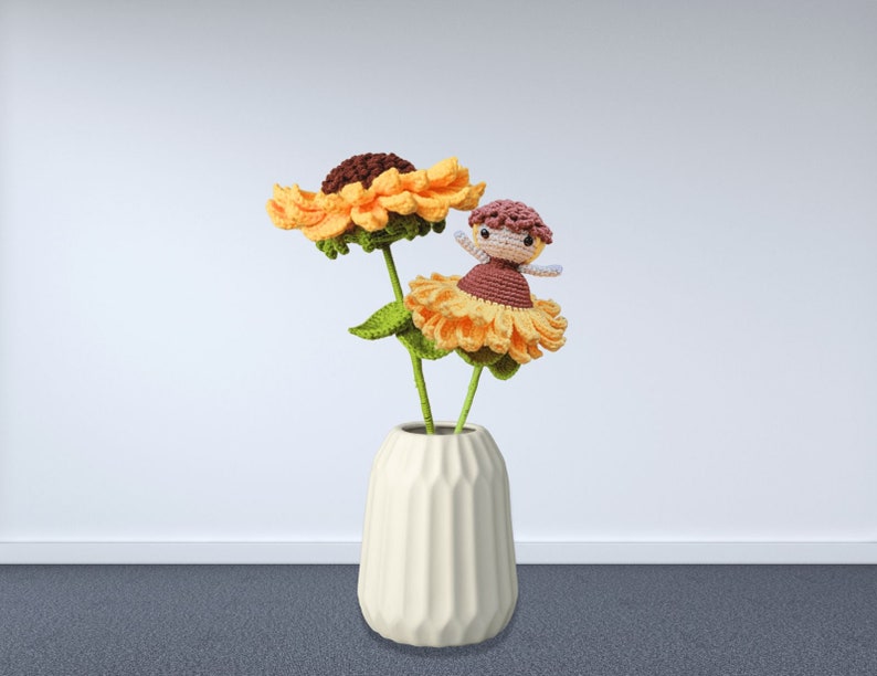 May include: A cream-colored vase holds two crocheted sunflowers with green stems and leaves. A small crocheted doll with a brown dress and a pink head is perched on one of the flowers. The sunflowers are yellow and brown.