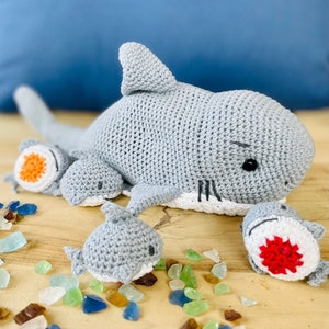 Puede incluir: Un conjunto de cuatro juguetes de tiburón de crochet gris. El tiburón más grande está acostado con la boca abierta. Los otros tres tiburones son más pequeños y están de pie. Los tiburones están hechos con un hilo suave y de felpa y tienen ojos negros y dientes blancos. Los tiburones están rodeados de pequeños trozos de vidrio de mar.