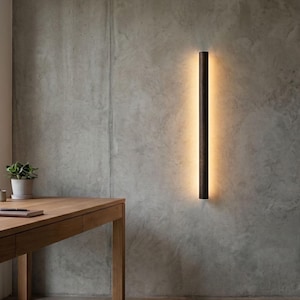 Op de afbeelding: Een modern interieur met een houten bureau, een mok, een notitieboek en een plant in een pot. Een lange, verticale wandlamp met een warme gloed is bevestigd aan een betonnen muur. De kamer is gevuld met natuurlijk licht.