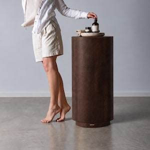 Può includere: Una camicia bianca abbottonata, una canotta beige e pantaloncini beige. Un piedistallo cilindrico marrone scuro si trova nelle vicinanze, sormontato da un piccolo vassoio in pietra contenente una bottiglia in vetro ambrato e un vasetto nero con etichetta.