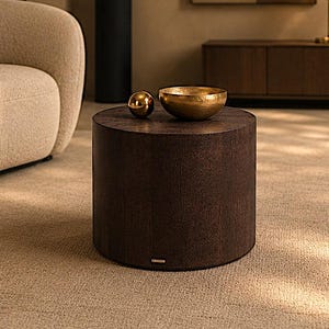Pode incluir: Uma mesa lateral cilíndrica de madeira castanho-escura com acabamento texturizado. Em cima da mesa encontra-se uma pequena esfera de ouro metálico e uma taça rasa de ouro martelado. A mesa está posicionada numa sala com um sofá bege.