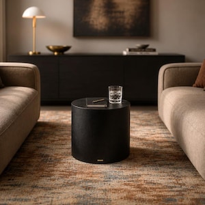 May include: A cylindrical black coffee table sits between two beige sofas on a textured rug with brown and gray patterns. On the table, there is a clear glass tumbler and a small notebook with a pen. The room features a dark sideboard and lamp.