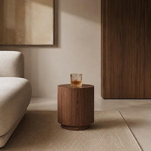 May include: A cylindrical wooden side table with a dark brown grain finish and a stepped base. A clear glass tumbler containing an amber-coloured liquid sits on the flat top surface of the table, which is positioned in a room with neutral decor.