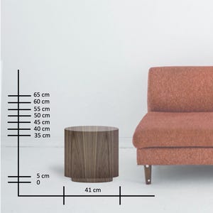 May include: A cylindrical wooden side table with a dark brown finish. The table is 41 cm wide and 40 cm tall. The table is next to a rust-colored sofa. The background is white.