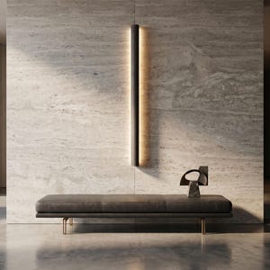 Puede incluir: Escena interior moderna con un banco largo gris oscuro con patas doradas, una lámpara vertical negra con un brillo cálido y una escultura decorativa. El fondo es una pared texturizada de color claro.