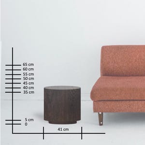 May include: A dark brown, cylindrical side table with a textured wood grain finish. The table is 16 inches in diameter and 26 inches tall. The base is slightly wider than the body. A rust-colored sofa is in the background.