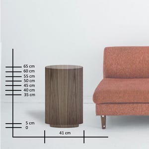 May include: A cylindrical wooden side table with a dark brown finish. The table is 25.6 inches tall and 16.1 inches wide. The table has a simple, modern design and is placed next to a coral-colored sofa.