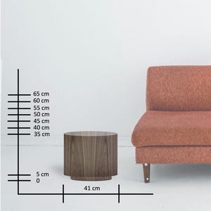 May include: A cylindrical wooden side table with a dark wood grain finish. The table is 16 inches wide and 26 inches tall. A rust-colored sofa is partially visible in the background.
