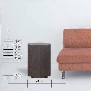 May include: A dark brown, cylindrical side table with a textured wood grain finish. The table is 25.6 inches tall and 16.1 inches wide. It has a slightly wider base and is positioned next to a rust-colored sofa.