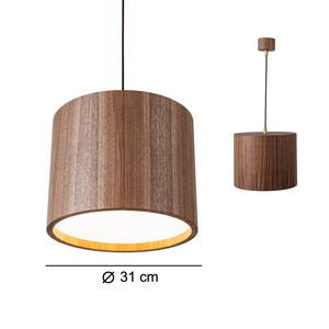 Puede incluir: Lámpara colgante de madera con una pantalla cilíndrica de 31 cm de diámetro, suspendida de un cable negro. La pantalla tiene un patrón de vetas de madera visible y emite una luz cálida. La montura del techo también es de madera.