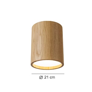 Könnte beinhalten: Zylindrische Holzleuchte mit einem Durchmesser von 21 cm. Die Leuchte hat eine natürliche Holzmaserung und ein warmes Leuchten, das von der offenen Unterseite ausgeht. Das Design ist schlicht und modern.