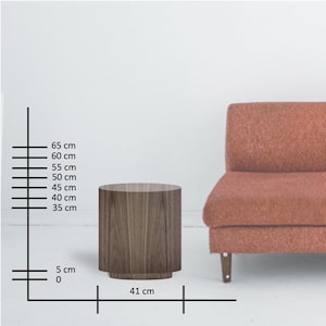 May include: A cylindrical wooden side table with a dark wood grain finish. The table is 16 inches in diameter and 26 inches tall. A reddish-orange sofa is partially visible in the background.