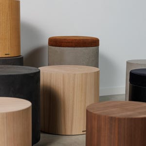 May include: A collection of cylindrical wooden stools in various shades of brown and gray. Some stools have a solid wood appearance, while others feature a contrasting upholstered top in a warm, earthy tone. The stools are arranged in a studio setting.