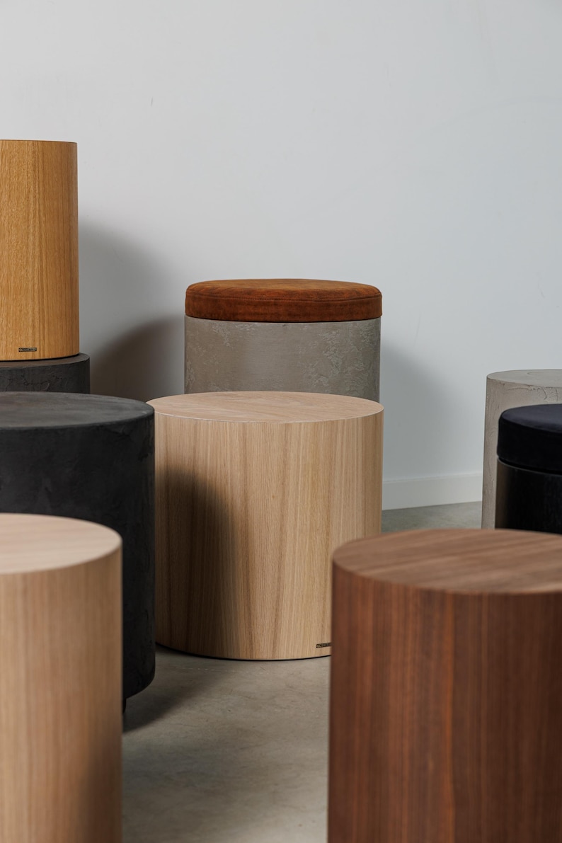 May include: A collection of cylindrical wooden stools in various shades of brown and gray. Some stools have a solid wood appearance, while others feature a contrasting upholstered top in a warm, earthy tone. The stools are arranged in a studio setting.