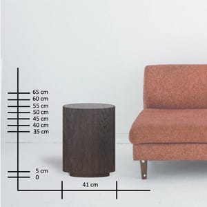 May include: A dark brown, cylindrical side table with a slightly flared base. The table is approximately 25.6 inches tall and 16.1 inches wide. The table is next to a rust-colored sofa.