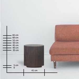 May include: A dark brown, cylindrical side table with a slightly flared base. The table is 41 cm wide and 65 cm tall. The table is next to a rust-colored sofa. The table is made of wood.