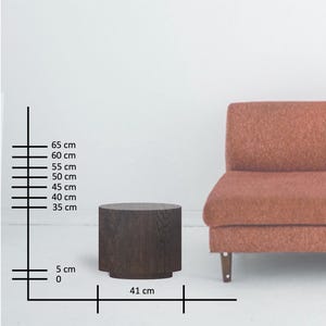 May include: A dark brown, cylindrical side table with a flat top and a slightly wider base. The table is 16 inches wide and 26 inches tall. A reddish-brown sofa is partially visible in the background.