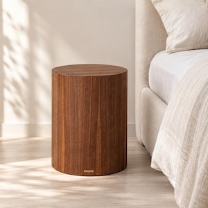 May include: A cylindrical side table crafted from dark brown wood with a visible vertical grain pattern. The table features a flat circular top and a small rectangular metallic brand plate attached near the base on the front side.