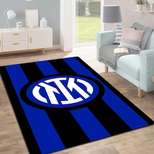 Könnte beinhalten: Ein rechteckiger Teppich mit blau-schwarzem Streifenmuster. In der Mitte befindet sich ein weiß-blaues Logo in einem Oval. Der Teppich liegt auf einem Holzboden in einem Wohnzimmer.