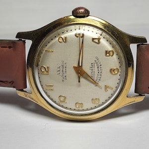 Puede incluir: Un reloj de pulsera automático AXA dorado vintage con esfera clara, números y agujas negros. El reloj tiene una correa de cuero marrón y el texto "AXA automatic 25 JEWELS" es visible en la esfera.