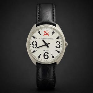 Peut inclure: Une montre à quartz Luc Desroches avec un boîtier argenté et un bracelet en cuir noir. Le cadran blanc présente des chiffres arabes noirs aux positions 3, 6 et 9, des index noirs, des aiguilles noires et un symbole marteau et faucille rouge.