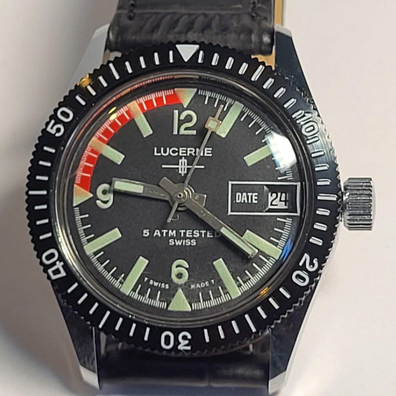 May include: A vintage Lucerne watch with a black dial, luminous hands and markers, and a date display. The watch has a black rotating bezel with white markings and a black leather strap. The watch is marked "5 ATM TESTED SWISS".