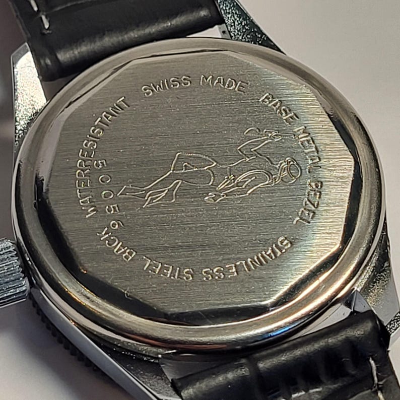 May include: Close-up of the back of a silver wristwatch with a black leather band. The watch back is engraved with a diver figure and the words "SWISS MADE", "WATER RESISTANT 50 M", "STAINLESS STEEL BACK", and "BASE METAL BEZEL".