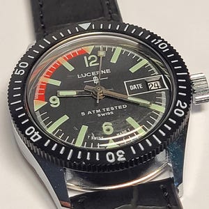May include: Close-up of a Lucerne watch with a black dial, luminous hands, and a date display. The watch has a black bezel with white markings and a black leather strap. The watch face displays the text "LUCEF.NE" and "5 ATM TESTED SWISS".