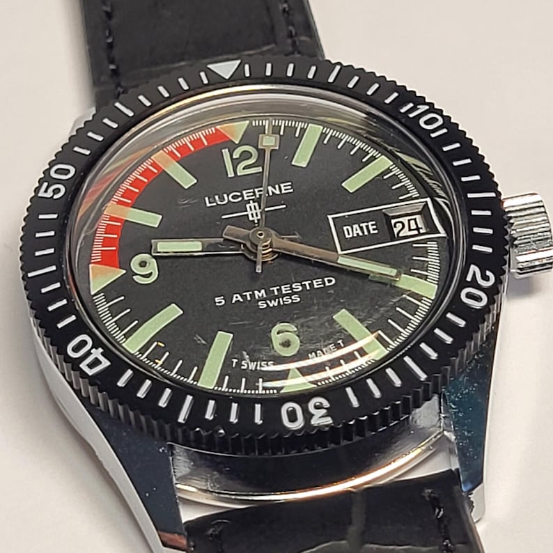 May include: Close-up of a Lucerne watch with a black dial, luminous hands, and a date display. The watch has a black bezel with white markings and a black leather strap. The watch face displays the text "LUCEF.NE" and "5 ATM TESTED SWISS".