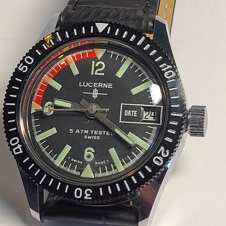 May include: A Lucerne watch with a black dial, luminous hands and markers, and a date display at the 3 o'clock position. The watch has a black rotating bezel and a black leather strap. The watch is marked "5 ATM TESTED SWISS".