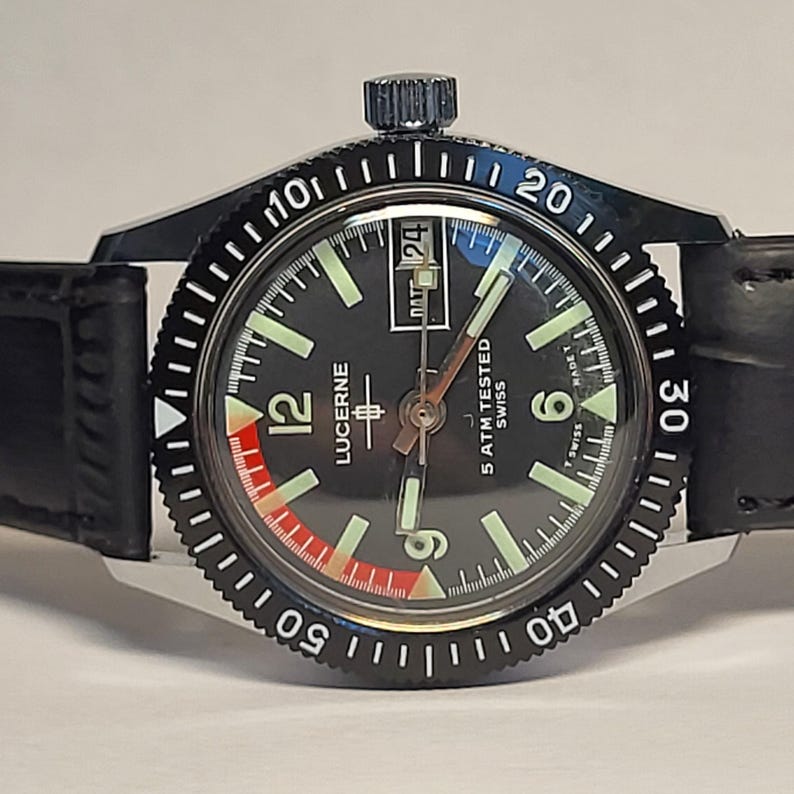 May include: A black Lucerne wristwatch with a black bezel and a black leather band. The watch face is black with white numerals and hands, and a red and white accent. The watch reads "5 ATM TESTED SWISS".
