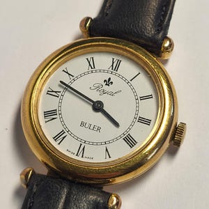 Könnte beinhalten: Eine goldfarbene Buler Royal Armbanduhr mit weißem Zifferblatt und schwarzen römischen Ziffern. Die Uhr hat ein schwarzes Lederarmband und eine kleine Krone. Das Zifferblatt zeigt die Wörter "Royal" und "Buler" sowie "Swiss Made".