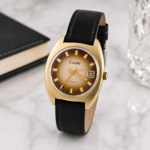 Puede incluir: Un reloj vintage dorado con una correa de cuero negro. La esfera del reloj tiene un degradado marrón y crema con manecillas y marcadores dorados. La fecha se muestra a las 3 en punto. El reloj está etiquetado como "Excelle".