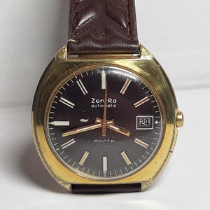 May include: A vintage gold-toned wristwatch with a dark brown face and a brown leather band. The watch face displays the text "Zen Ra automatic" and "MANTA", with a date window at the 3 o'clock position. The watch has gold hands and hour markers.