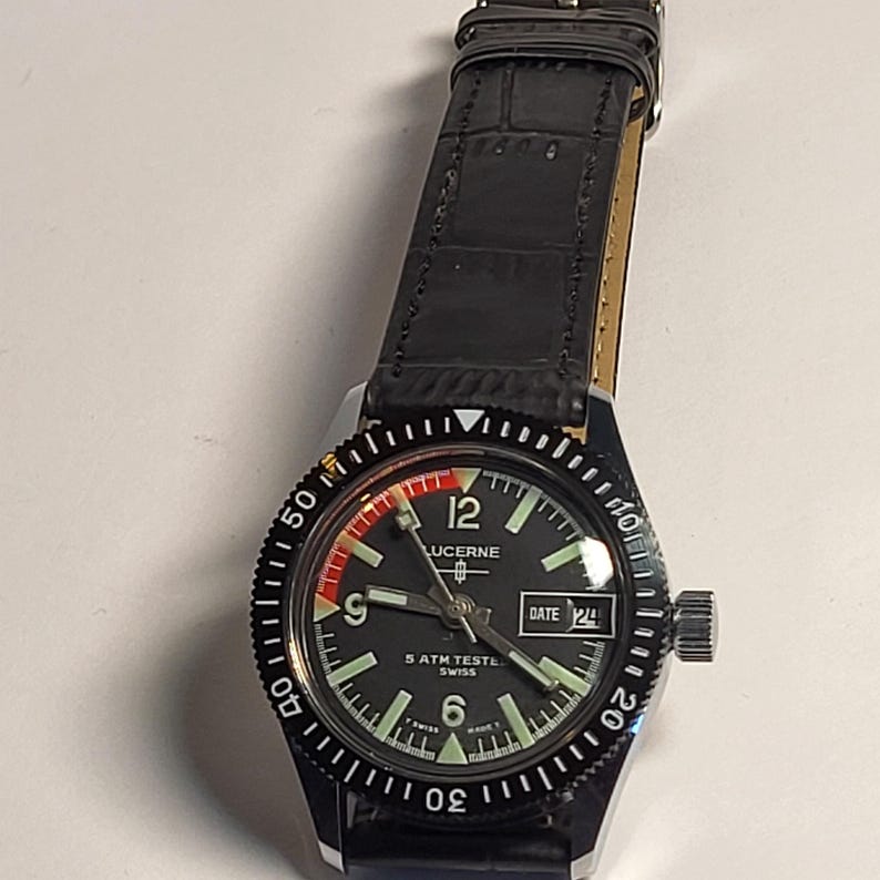 May include: Black Lucerne watch with a black bezel and a black leather band. The watch face is black with white numerals and hands. The watch has a date display at the 3 o'clock position. The watch is marked "5 ATM TESTED SWISS".