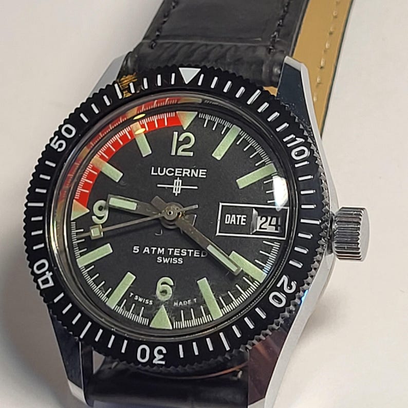 May include: A Lucerne wristwatch with a black dial, luminous hands and markers, and a date display at the 3 o'clock position. The watch has a black rotating bezel and a black leather strap. The watch face displays the text "LUCERNE" and "5 ATM TESTED SWISS".