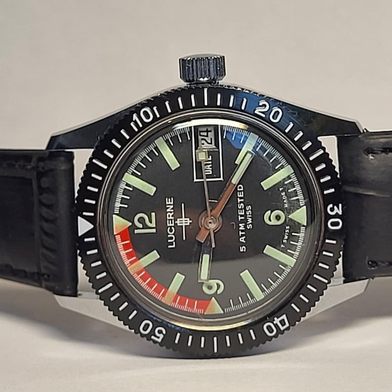 May include: A black Lucerne watch with a black bezel and a black leather strap. The watch face is black with white and red accents, luminous hands, and a date display. The watch is marked "5 ATM TESTED SWISS".