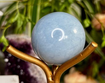 May include: A light blue crystal sphere, approximately 2 inches in diameter, displayed on a gold-colored metal stand. The sphere has a smooth, polished surface. Green foliage is visible in the background, adding a natural element to the composition.