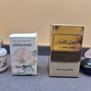 Könnte beinhalten: Auf einem Tablett werden vier Parfümflaschen und -schachteln präsentiert. Zu den Flaschen gehören eine weiße Flasche mit silbernem Verschluss, eine blaue Flasche mit rosa Oberteil und zwei Parfümkartons. Die Kartons sind mit "Anais Anais" und "Parfum de Nuit" beschriftet.