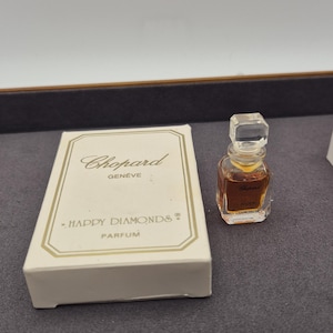 Könnte beinhalten: Eine weiße Schachtel mit goldenen Schriftzügen, auf der "Chopard Genève" und "Happy Diamonds Parfum" steht. Neben der Schachtel befindet sich ein kleines, klares Glasfläschchen mit Parfum, auf dem in goldenen Buchstaben "Chopard Happy Diamonds" steht.