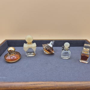 Könnte beinhalten: Fünf Miniatur-Parfümflacons in einem Holztablett mit grauer Filz-Auskleidung. Die Flacons haben verschiedene Formen und Größen und sind mit goldenen und silbernen Akzenten verziert. Die Flacons enthalten eine klare Flüssigkeit.