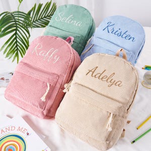 May include: Four corduroy backpacks in pastel colors: pink, mint green, light blue, and beige. Each backpack has a front pocket and a name embroidered on the front: Raffy, Selina, Kristen, and Adelya. The backpacks are on a white surface.