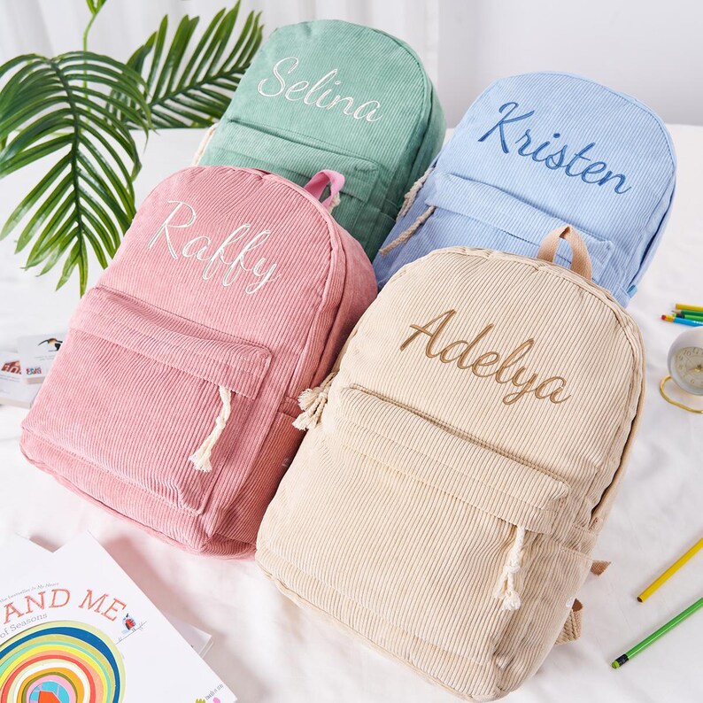 May include: Four corduroy backpacks in pastel colors: pink, mint green, light blue, and beige. Each backpack has a front pocket and a name embroidered on the front: Raffy, Selina, Kristen, and Adelya. The backpacks are on a white surface.