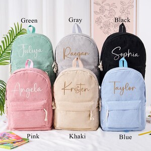 May include: A collection of corduroy backpacks in various colors: green, gray, black, pink, khaki, and blue. Each backpack features a name embroidered on the front. The backpacks have a front pocket and a top handle.