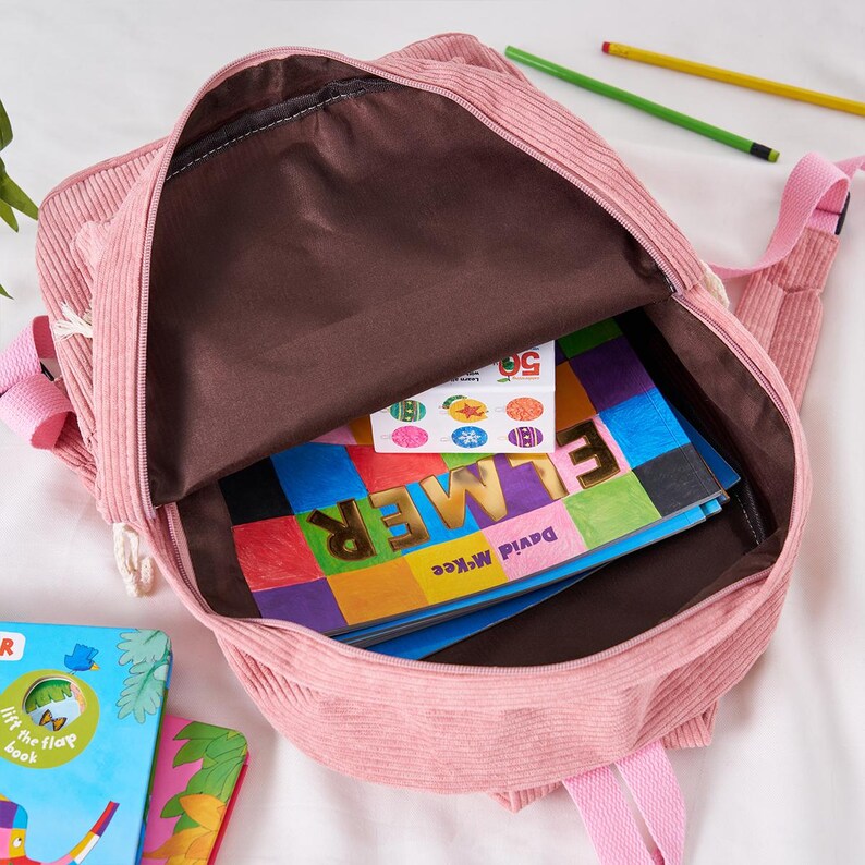 May include: A pink corduroy backpack with an open zipper, revealing a brown interior and colorful books. The backpack has pink straps and white rope detailing. Books include the title "Elmer" by David McKee.