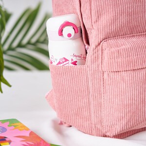 May include: A pink corduroy backpack with a white and pink insulated water bottle in the side pocket. The water bottle has a pink lid and the words "Sweet Heart" printed on it, along with a cake and heart design.
