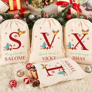 May include: Four personalised Christmas gift bags with red and green designs. The bags are made of natural fabric and have a drawstring closure. Each bag features a large red letter, a small Santa Claus figure, and a personalised name. The names on the bags are Salome, Vicky, Xmas, and Teddy.