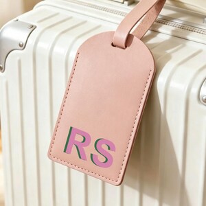 以下が含まれることがあります： 上部が丸みを帯びたペールピンクのラゲッジタグ。同色のストラップが付いており、緑とピンクの重なり合う太いフォントで「RS」の文字がプリントされています。タグは縦に溝が入った白いハードシェルタイプのスーツケースに取り付けられています。