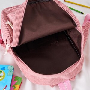 May include: Open view of a pink corduroy backpack with a brown interior. The backpack has a front pocket with a drawstring closure and pink straps. Pencils and a book are visible in the background.