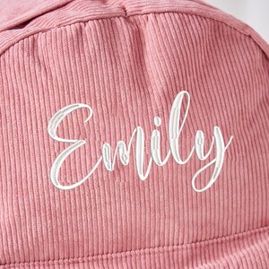 May include: Close-up of a dusty rose corduroy fabric with the name "Emily" embroidered in white cursive script. The fabric has vertical ridges, and the embroidery is centered.